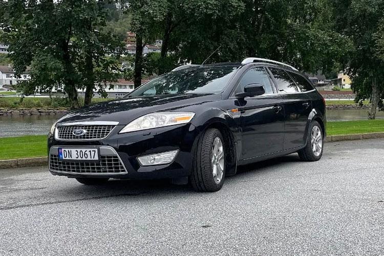 En svart Ford stasjonsvogn parkert ved elven, med trær i bakgrunnen.