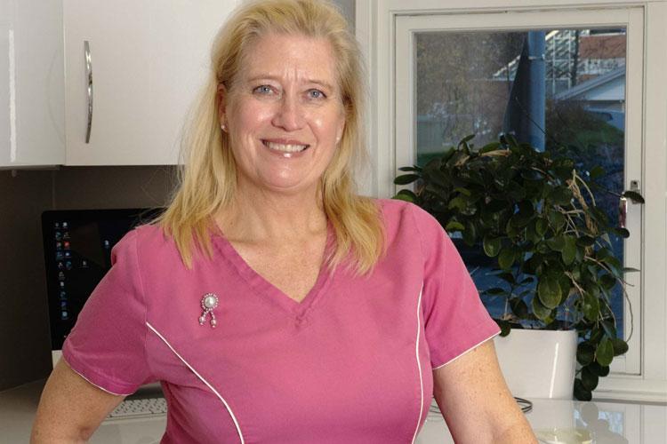 Kvinne i rosa arbeidsdrakt smiler foran et vindu med planter og en datamaskin i bakgrunnen.
