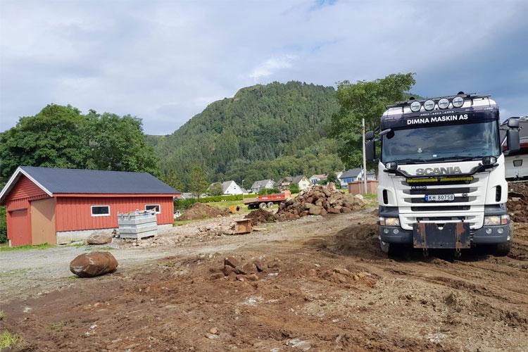 En lastebil fra Scania står på en byggeplass med en rød hytte og grønne fjell i bakgrunnen.