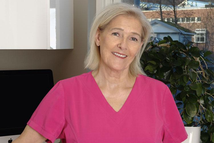 En smilende kvinne i rosa arbeidsdrakt står foran en datamaskin og vindu med planter i bakgrunnen.