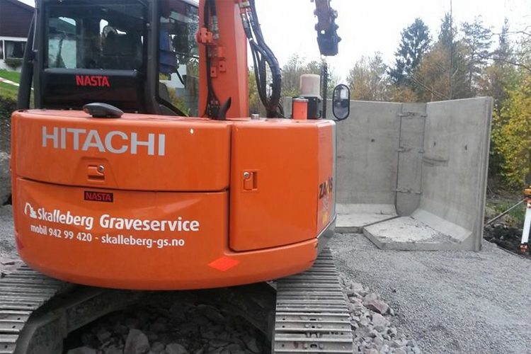 En oransje gravemaskin fra Hitachi ved en byggeplass med betongvegg i bakgrunnen.