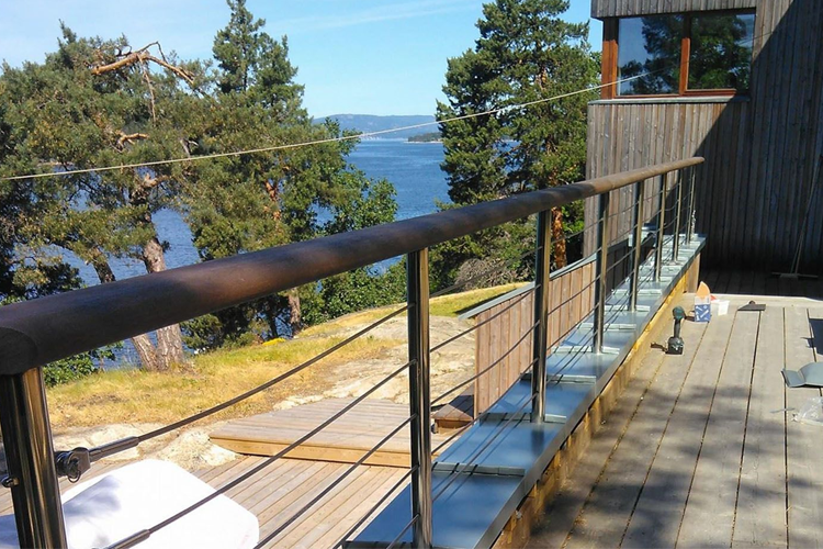 Utsikt over vannet fra en moderne terrasse med rekkverk av tre og stål, omringet av trær.