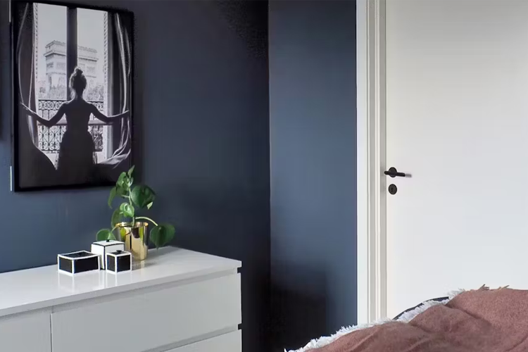 Soverom med mørk vegg, hvit kommode, plante og kunstverk av en silhuett ved vinduet.