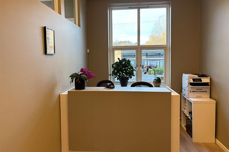 Moderne resepsjon med planter, vindu og kontormaskiner i et lyst rom.