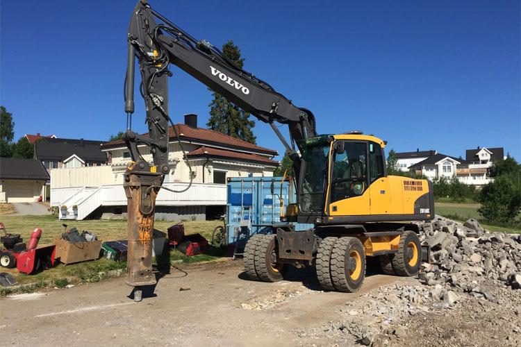 Gravemaskin fra Volvo med hydraulisk hammer på byggeplass, omgitt av byggeutstyr og boliger i bakgrunnen.