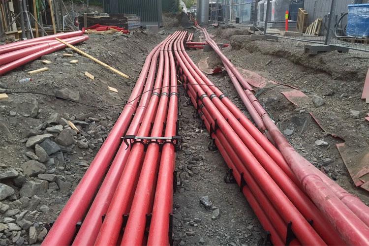 Røde rør lagt i en grusgrav på en byggeplass, med jord og byggeutstyr i bakgrunnen.