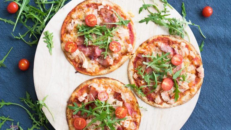 Tre pizzaer med bacon, tomater og ruccola på en treplate, omringet av friske urter.