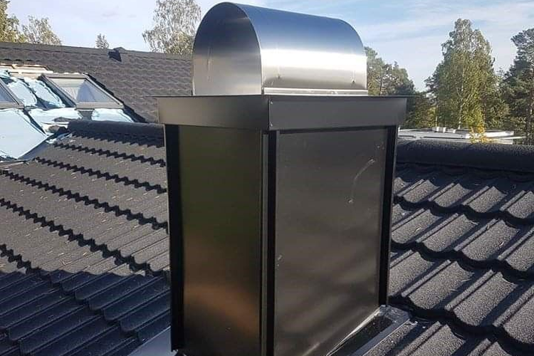 Kraftig takventilasjon med metallhette på et tak med takstein og trær i bakgrunnen.