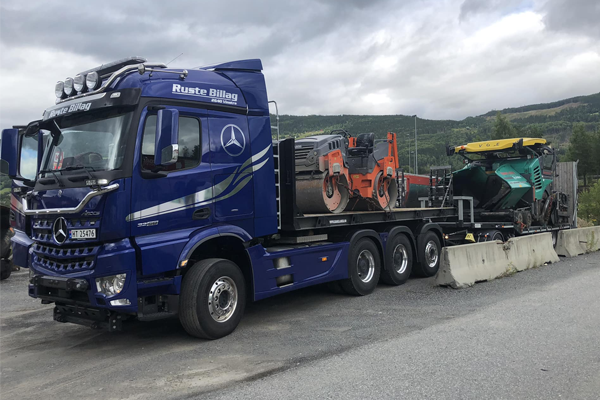 En blå lastebil fra Mercedes-Benz med to maskiner på tilhenger, parkert ved en grå betongbarriere.