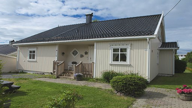 En koselig enebolig med terrasse, hage og grusvei under en blå himmel med skyer.