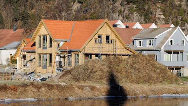 Moderne hus med rød takstein og treverk, bygget ved vannkanten med landskap i bakgrunnen.