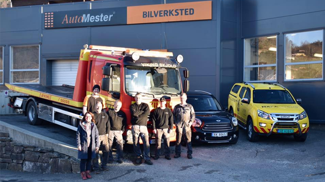 En gruppe ansatte står foran et bilverksted med en lastebil og to biler parkert.