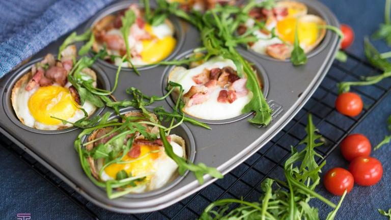 Egg med bacon og ruccola i muffinsform, servert med cherrytomater på en blå bakgrunn.