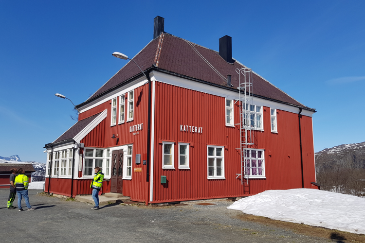 Rødt hus med hvite vinduer og tak, omgitt av snø og fjell, med personer i arbeidstøy i nærheten.