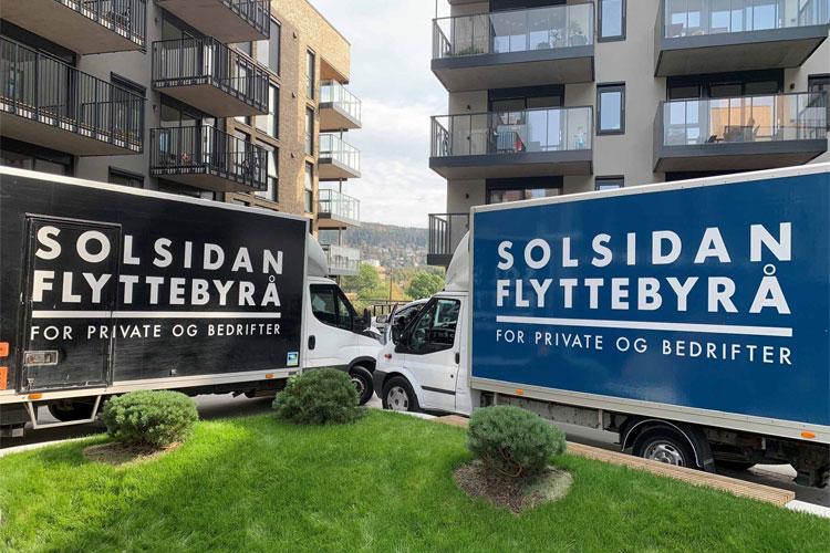 To lastebiler med påskriften "Solsidan Flyttebyrå" parkert ved en bygning, med grønt gress foran.