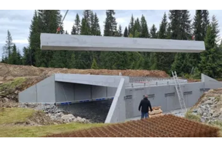 Betongplater heises over en byggeplass med en tunnel under konstruksjon, omgitt av trær og natur.