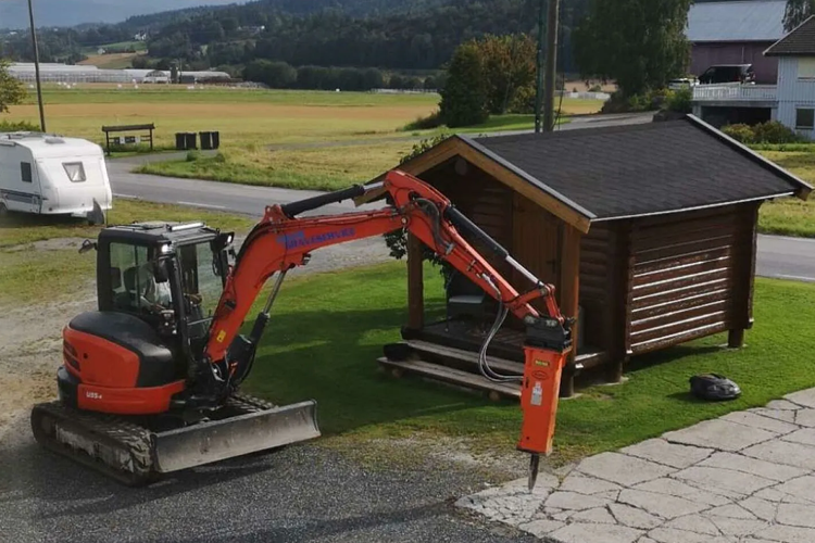 Gravemaskin med hydraulisk hammer står ved en hytte på en grønn plen i et landlig område.