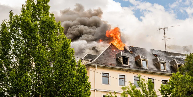 Brann på taket av en bygning med svart røyk og flammer som stiger opp mot himmelen. Grønn tre i forgrunnen.