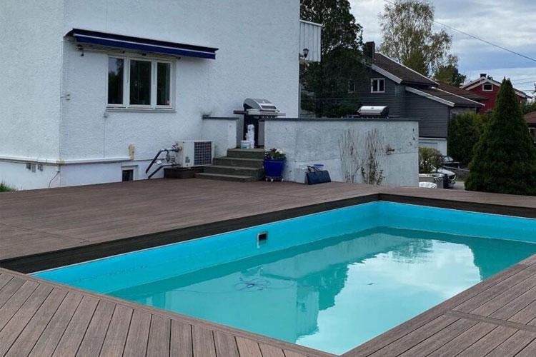 En hage med en svømmebasseng, tredekk og en hvit husvegg i bakgrunnen.