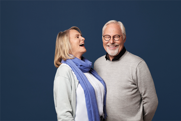 Et smilende eldre par står sammen mot en blå bakgrunn, med kvinnen som ler og mannen som smiler.