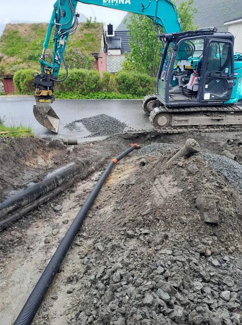 Gravemaskin som arbeider på en byggeplass med grus og rør synlig i bakken.