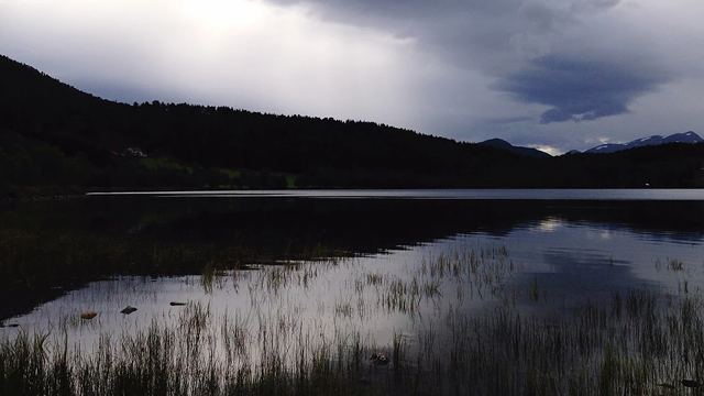 En stille innsjø med mørke skyer og fjell i bakgrunnen, omkranset av grønt gress og siv.