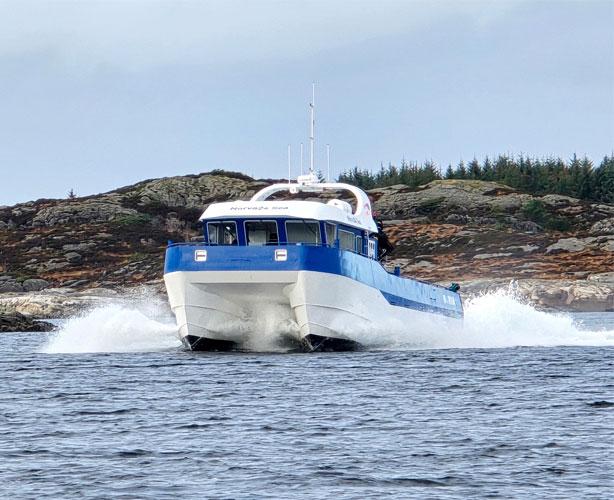 En hurtigbåt med blå og hvit farge som kjører gjennom vannet, omgitt av steinete kystlinje og trær.