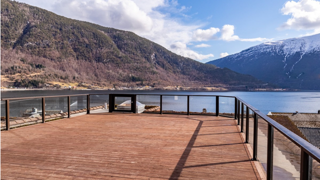 Utsikt fra en terrasse med tredekk, fjell og vann i bakgrunnen under en klar himmel.