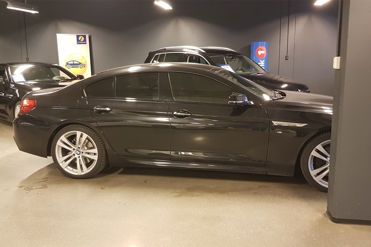 En svart bil med mørke vinduer står parkert i en garasje med flere andre biler i bakgrunnen.