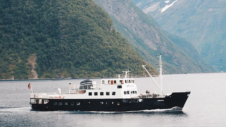 En svart og hvit ferge seiler gjennom fjorden med fjell i bakgrunnen.