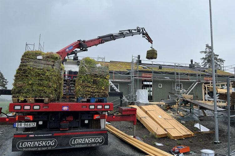 Lastebil med kran som løfter en stor kasse med materialer til taket på en bygning under konstruksjon.