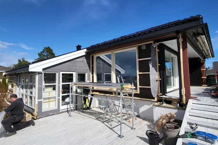 Hus med moderne arkitektur, arbeidere som jobber på fasaden, og solfylt himmel i bakgrunnen.