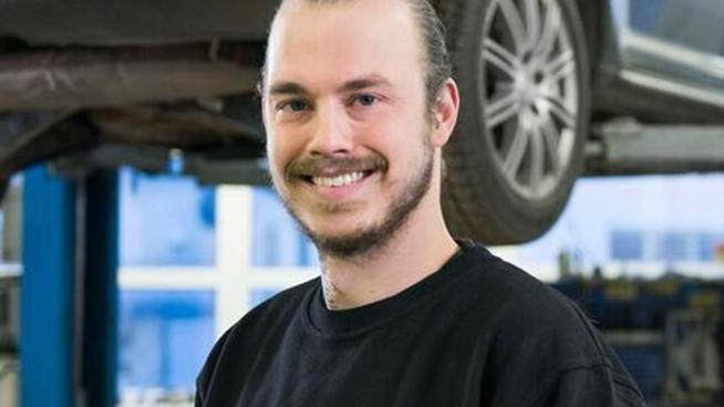 En smilende mekaniker i verkstedet, iført svart genser, står foran en hevet bil med verktøy i hånden.