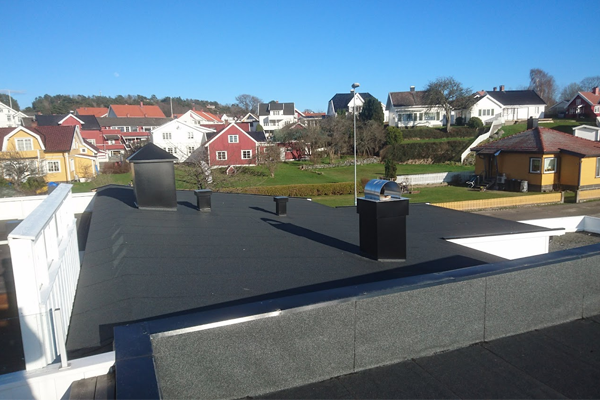 Utsikt fra taket med svarte takplater, skorsteiner og hus i bakgrunnen under en klar blå himmel.