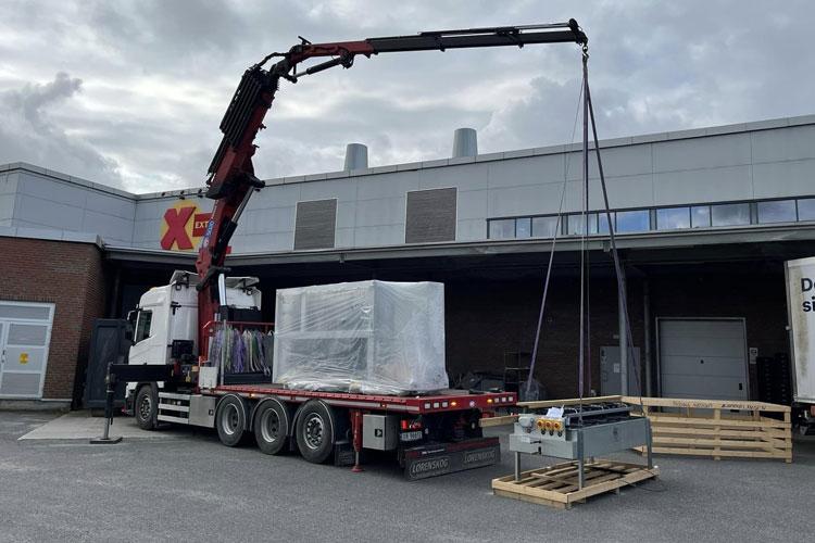 Lastebil med kran som løfter en stor pakke ved en bygning med X-logo. Pallet står på bakken.