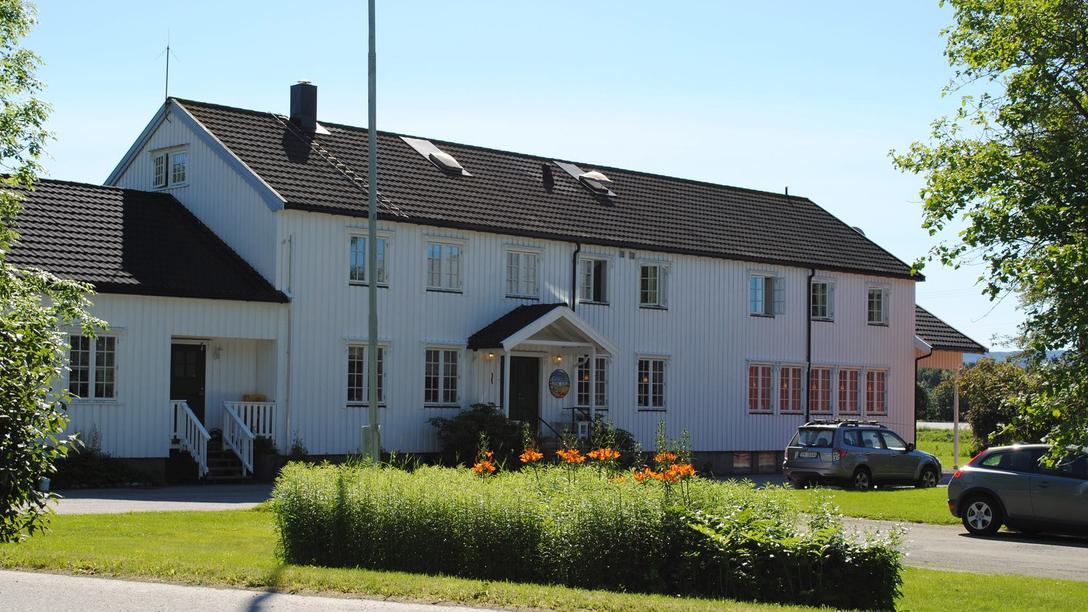 En stor, hvit bygning med svart tak, omgitt av grønt gress og blomster, med en bil parkert foran.