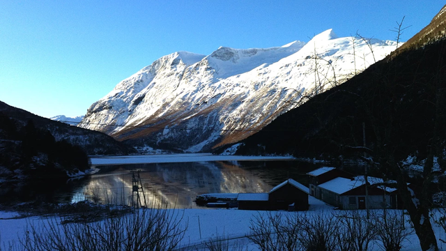 Vinterlandskap med snødekte fjell og speilbilde i innsjøen, med hytter i forgrunnen.