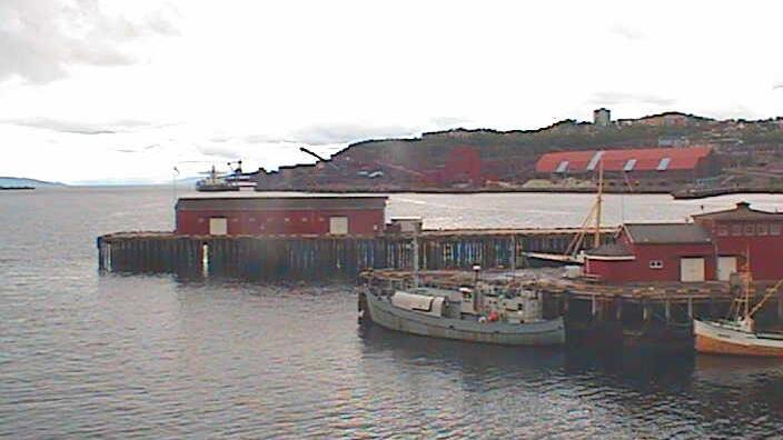 Bilde av en havn med røde bygninger, båter ved kaien og en stor båt i bakgrunnen under en skyet himmel.