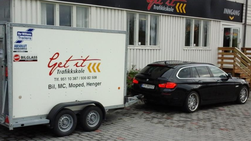 En bil med tilhenger parkert foran en trafikkskole med logo og informasjon på tilhengeren.