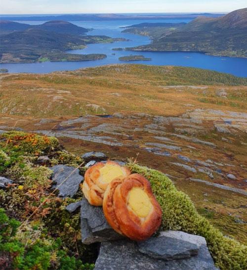 To brødskiver med ost ligger på steiner med utsikt over en vakker innsjø og fjellandskap.