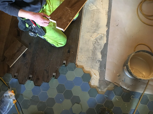 En håndverker legger gulvplanker på et hexagonalt flisgulv med lim i en romslig innendørs setting.