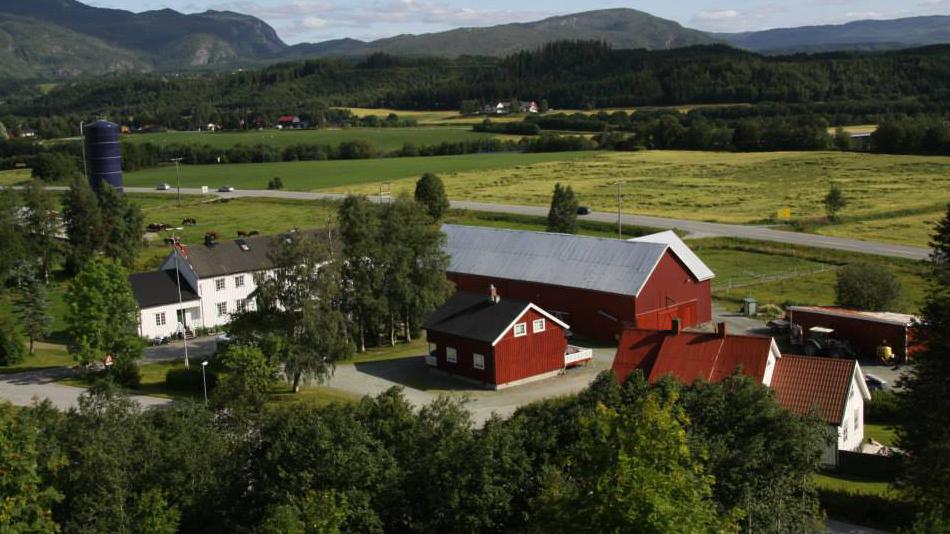 En vakker gård med røde bygninger, grønne åser og åkerlandskap under en blå himmel.