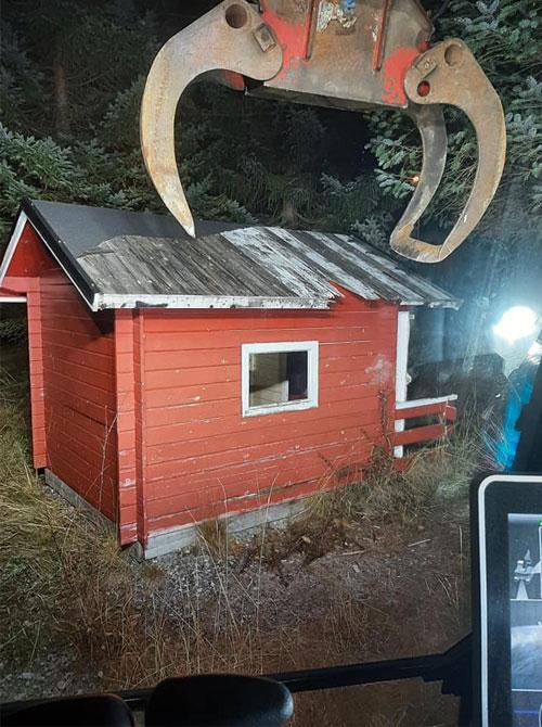 Rød hytte med skråtak i skogen, sett fra en maskin med gripere som løfter den.