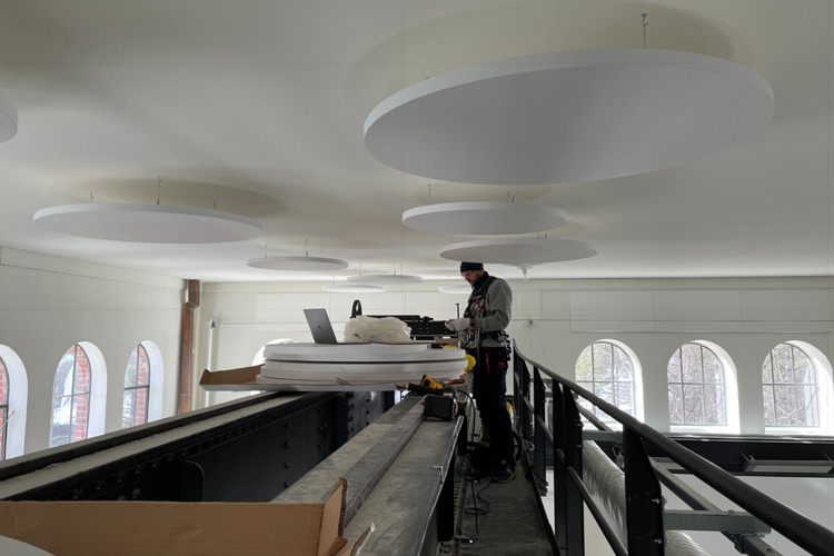 En arbeider står på en plattform med datamaskin, under store, runde taklamper i et rom med hvite vegger.