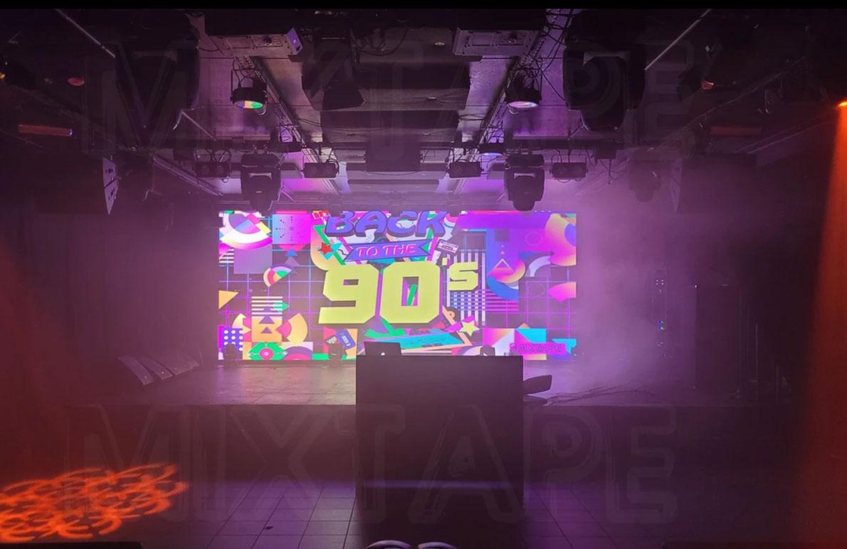 En fargerik scene med en stor skjerm som viser "90's" og et DJ-bord foran i et festlokale.