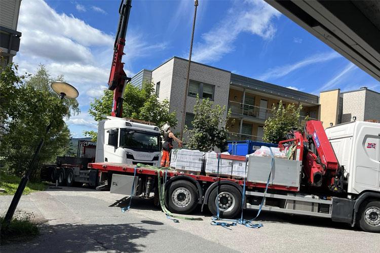 Lastebil med kran som laster av materialer ved en bygning på en solfylt dag.
