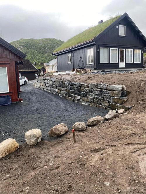 Moderne svart hus med gressdekt tak, steinmur og gruslagt innkjørsel i naturskjønt område.