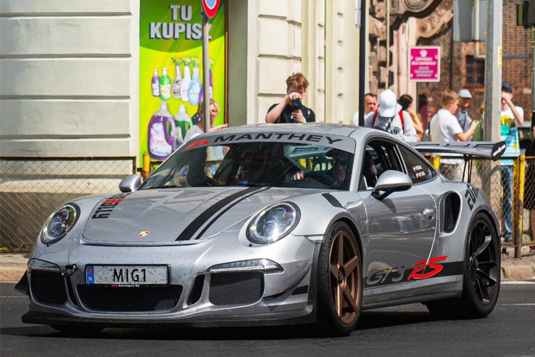 En sølvfarget Porsche 911 GT3 RS med racingdekor kjører forbi en folkemengde på gaten.