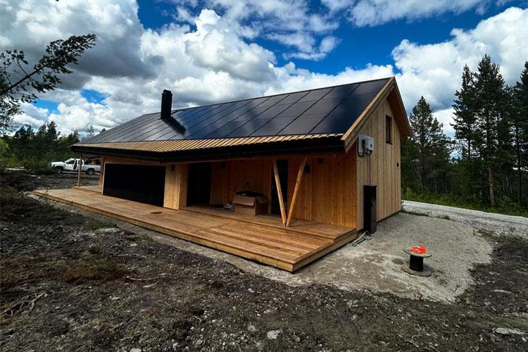 Moderne hytte med solcellepaneler, tredekk og terrasse, omringet av skog og blå himmel med skyer.
