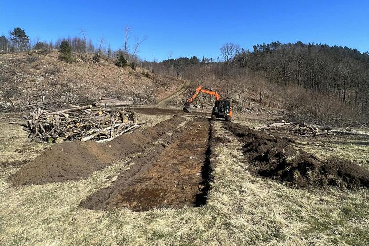 Gravemaskin arbeider på en åpen slette, med hogde trær og en klar blå himmel i bakgrunnen.